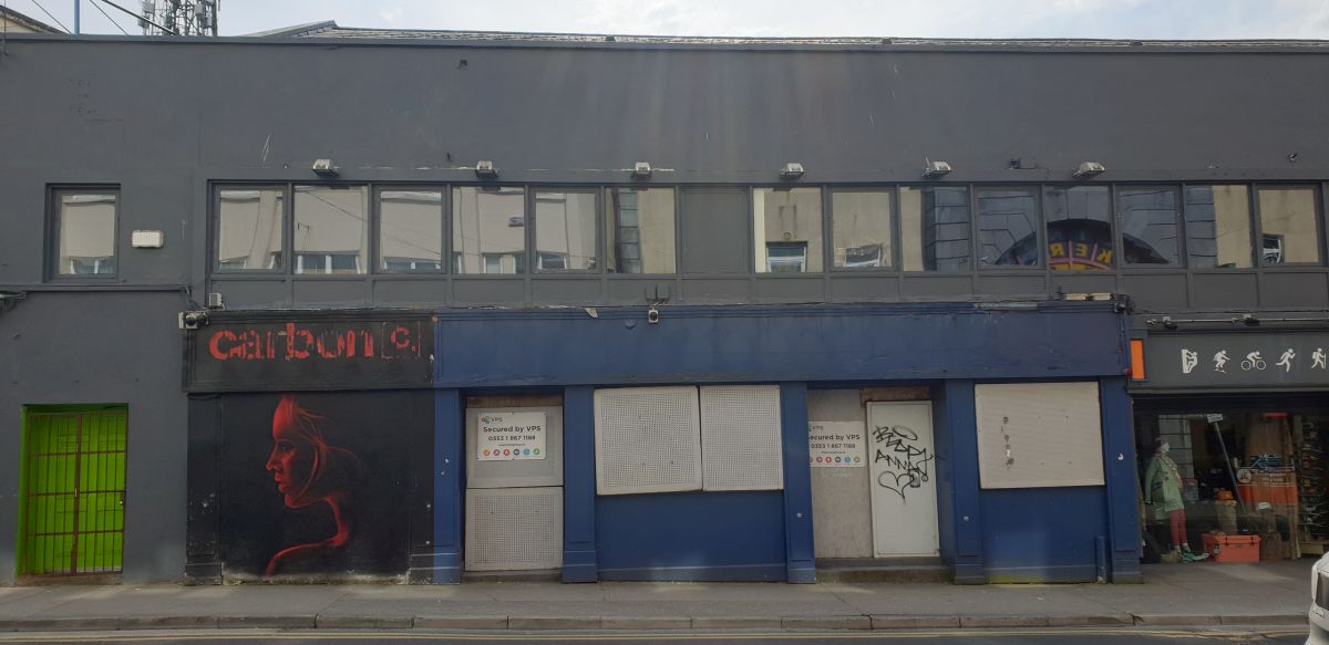 Two storey boarded up nightclub. Dark blue walls. Street art of a woman's face in profile in red and black on one section.